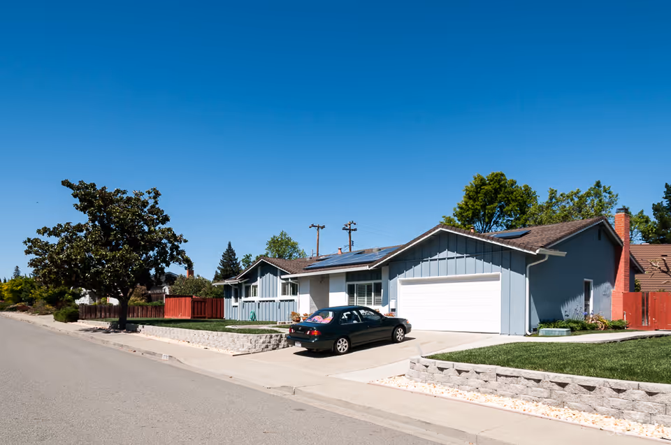 Single-story blue suburban house with a driveway, parked car, front lawn and clear blue sky.