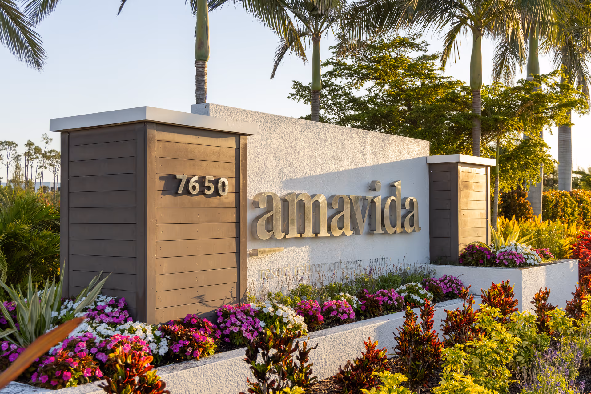 Stone and wood entrance sign reading 'amavida' with the number 7650, surrounded by colorful flowers and palm trees.