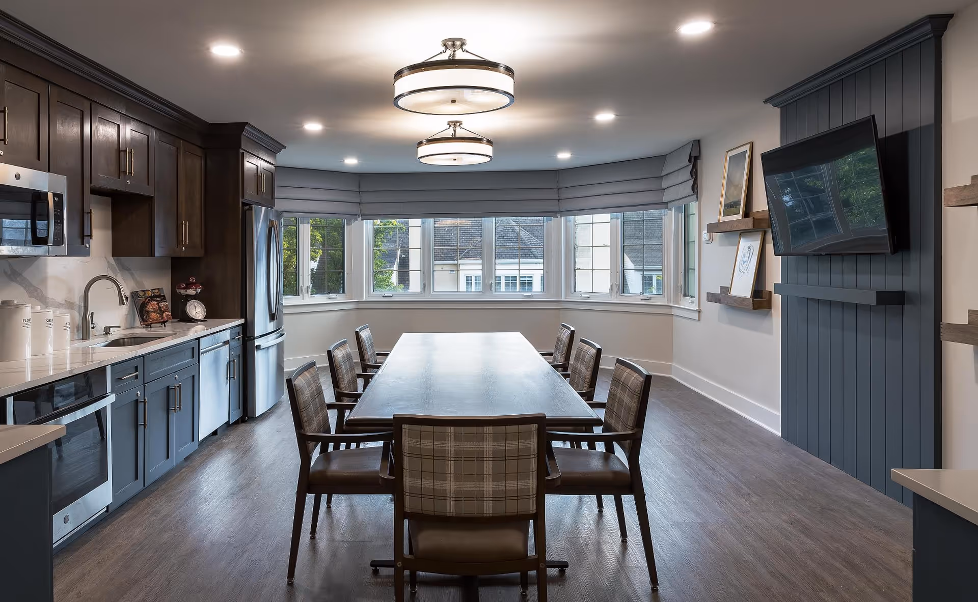 A modern kitchen and dining area with dark wood cabinets, stainless steel appliances including a microwave, oven, dishwasher, and refrigerator. A long wooden dining table with six plaid upholstered chairs is centered in the room. Large windows with gray Roman shades allow natural light to fill the space. Two round ceiling lights illuminate the room, and a flat-screen TV is mounted on a dark blue paneled wall with floating shelves displaying framed artwork.