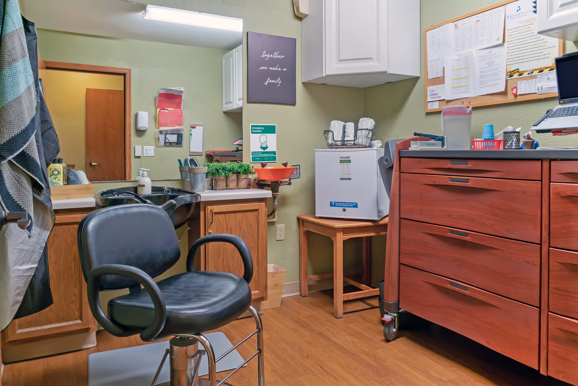 Interior view of a small salon or grooming room with a black salon chair in front of a black sink. The room has wooden cabinets, a small white refrigerator on a wooden stool, a bulletin board with papers pinned, and a wall sign that reads 'together we make a family'. There are various grooming supplies and hand sanitizer on the counters.