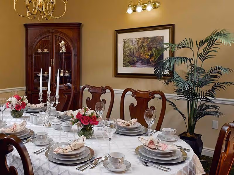 A formal dining room with a table set for six people. The table is covered with a white tablecloth and set with plates, bowls, cups, silverware, wine glasses, and cloth napkins. There are two floral centerpieces with pink and white flowers and two tall white candles in silver candle holders. Behind the table is a wooden china cabinet with glass doors displaying china, a framed nature photograph on the wall, and a large potted plant in the corner.