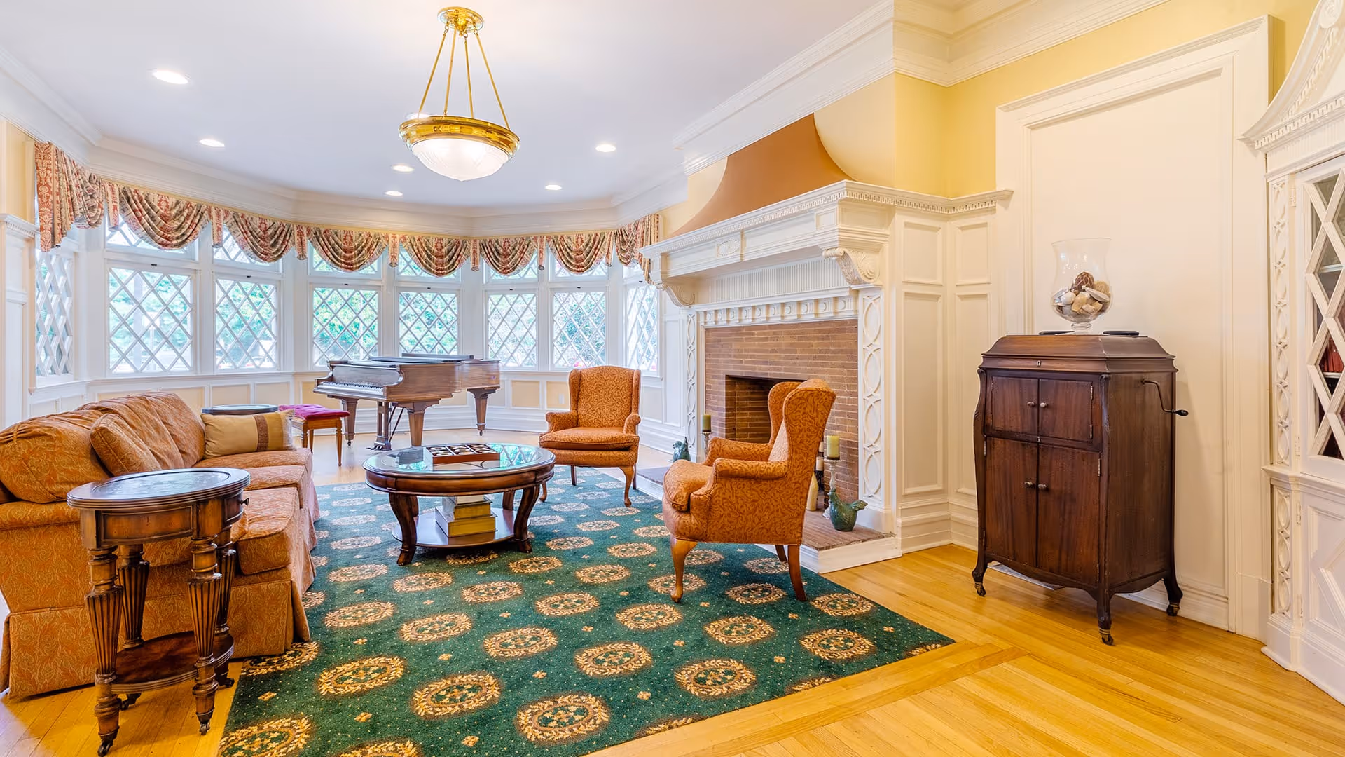 A bright and elegant living room with a green patterned carpet, an orange sofa, two matching armchairs, a round wooden coffee table, and a grand piano near a large bay window with decorative curtains. The room features a white ornate fireplace and wooden flooring.