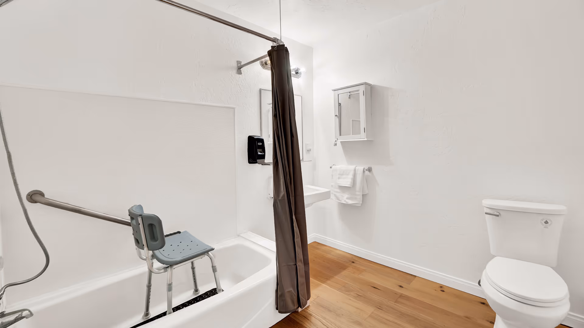 A clean and accessible bathroom with white walls and wooden floor. It features a bathtub with a gray shower chair and a grab bar, a brown shower curtain, a white toilet, a wall-mounted sink with a mirror above it, a towel rack with white towels, and a small white medicine cabinet.