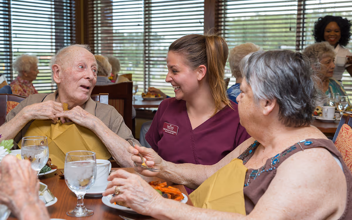 A group of elderly residents and a caregiver are seated around a dining table in a senior living facility. The caregiver, wearing a maroon uniform, is smiling and engaging warmly with two elderly residents who have yellow napkins tucked into their collars. Plates of food and glasses of water are on the table, with other residents and staff visible in the background near large windows with blinds.