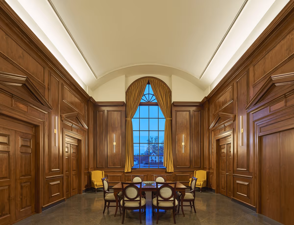 A formal wood-paneled common room with a round dining table and chairs beneath a tall arched window and high ceiling.