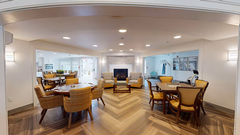 A spacious common area in a senior living facility featuring a cozy seating arrangement with two armchairs and a sofa around a fireplace. There are two tables with chairs, one with a chessboard set up. The room has wood-patterned flooring and is well-lit with recessed ceiling lights. Adjacent rooms with additional seating and exercise equipment are visible through open doorways.