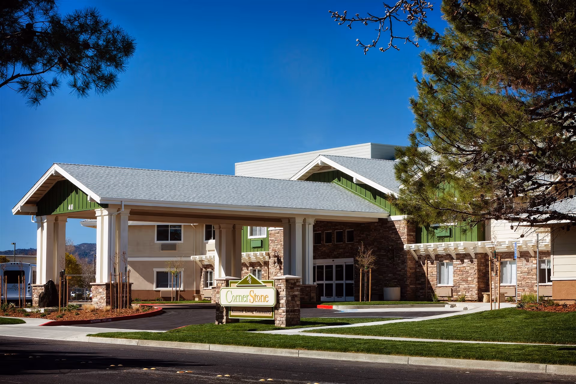 Exterior view of Cornerstone Assisted Living facility showing the main entrance with a covered drop-off area, green and beige building facade, stone accents, and a sign reading 'CornerStone Assisted Living Community' on a well-maintained lawn under a clear blue sky.