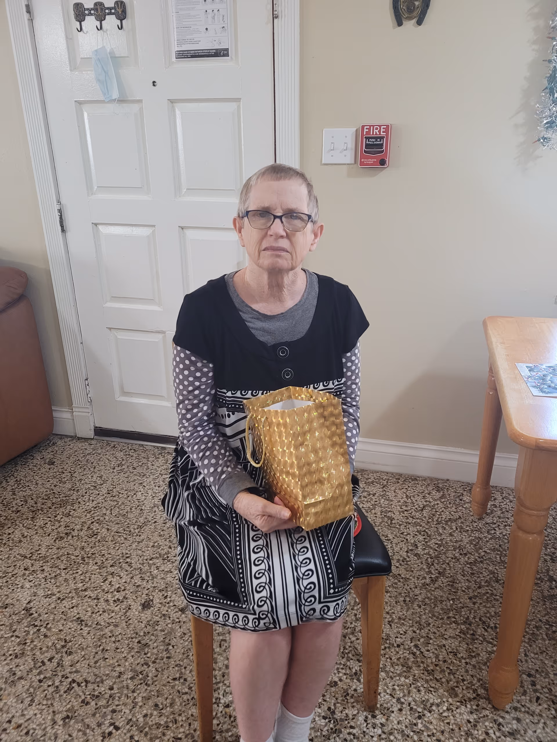 An elderly woman with short hair and glasses sitting on a chair indoors, holding a gold gift bag. She is wearing a black and white patterned dress with a gray long-sleeve shirt underneath. Behind her is a white door with a face mask hanging on a hook, a fire alarm on the wall, and part of a wooden table is visible to the right.