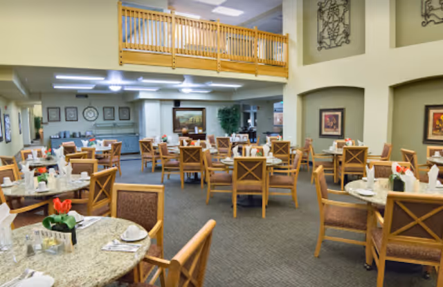 A spacious dining room with multiple round tables set with white napkins, cups, and small flower arrangements. Wooden chairs with cushioned seats surround the tables. The room features a high ceiling with a wooden balcony railing above, framed artwork on the walls, and soft lighting.
