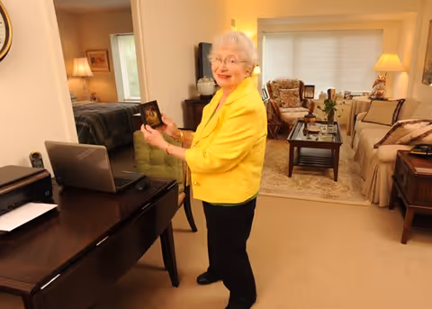 An elderly woman wearing a bright yellow jacket stands in a warmly lit living room holding a framed photo. The room features a beige carpet, a sofa with cushions, a coffee table with a plant, a lamp, and an armchair. A desk with a laptop and printer is visible in the foreground, and a bedroom can be seen through an open doorway.