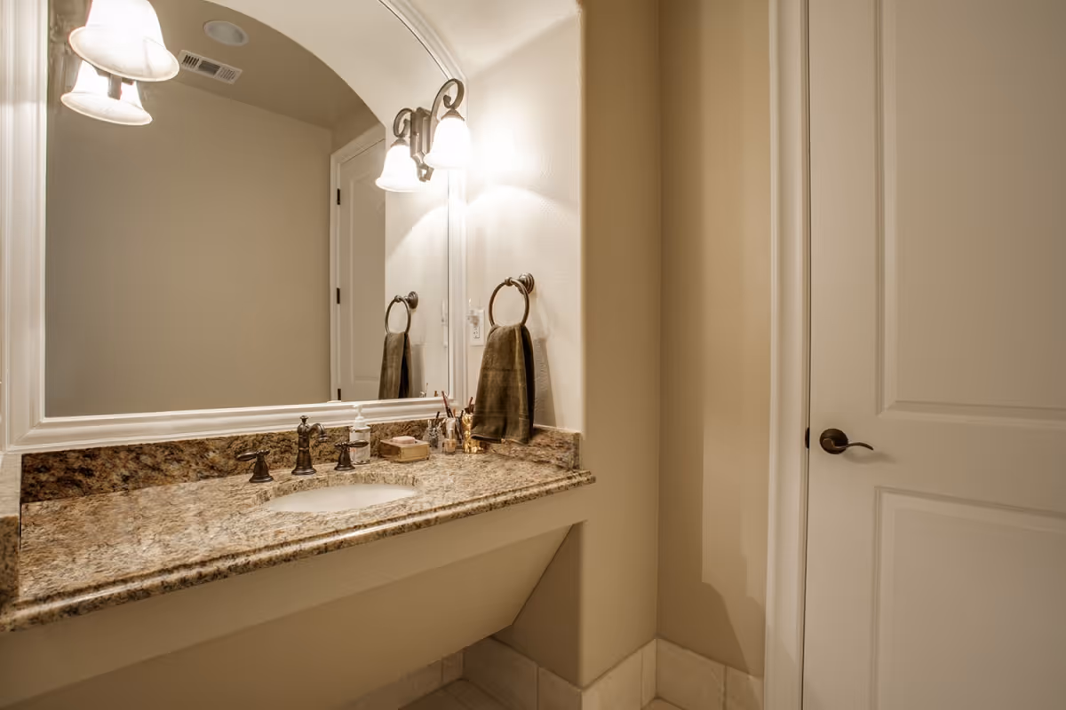 A bathroom vanity area with a granite countertop, an under-mount sink, bronze faucet fixtures, a large mirror with a white frame, two wall-mounted light fixtures with frosted glass shades, a towel ring holding a brown towel, and a closed white door with a bronze handle.