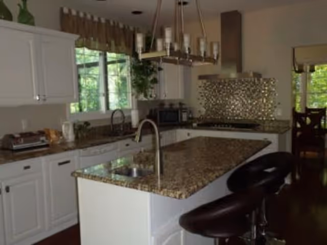 A modern kitchen with white cabinets, granite countertops, a kitchen island with a sink and two dark bar stools. There is a window above the sink with a valance, a decorative backsplash behind the stove, and a hanging light fixture above the island.