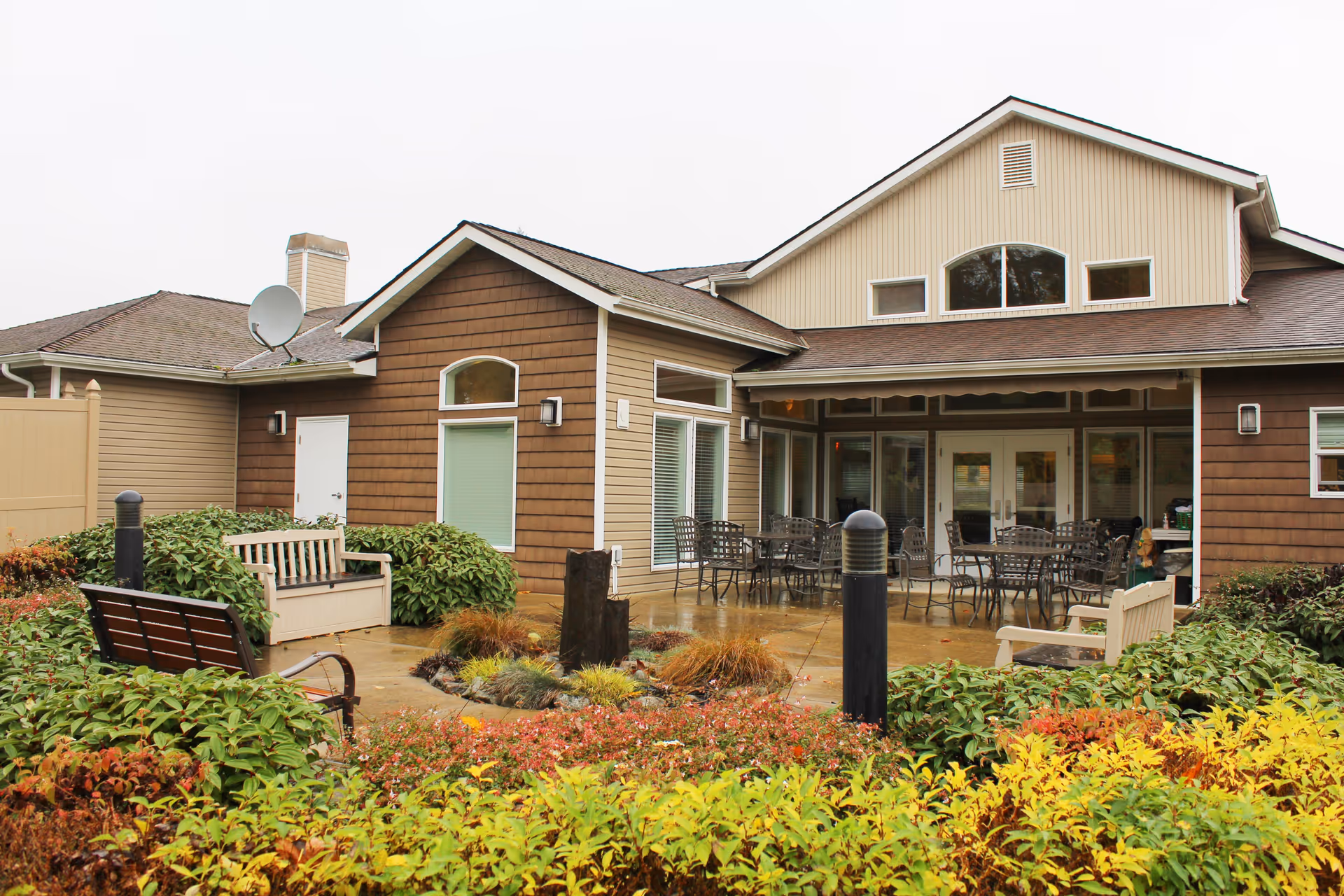Outdoor patio area at Normandy Park Senior Living featuring multiple metal tables and chairs under a covered section, surrounded by bushes and colorful plants. The building has beige and brown siding with several windows and a satellite dish on the roof.