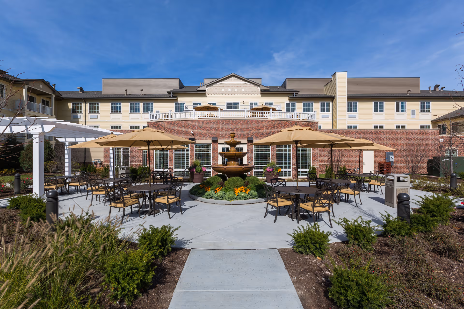 Outdoor patio area at Green Oaks Senior Living featuring multiple round tables with chairs and beige umbrellas. A three-tiered water fountain is centered among flower beds with colorful flowers and greenery. The building facade behind is a combination of brick and light-colored siding under a clear blue sky.