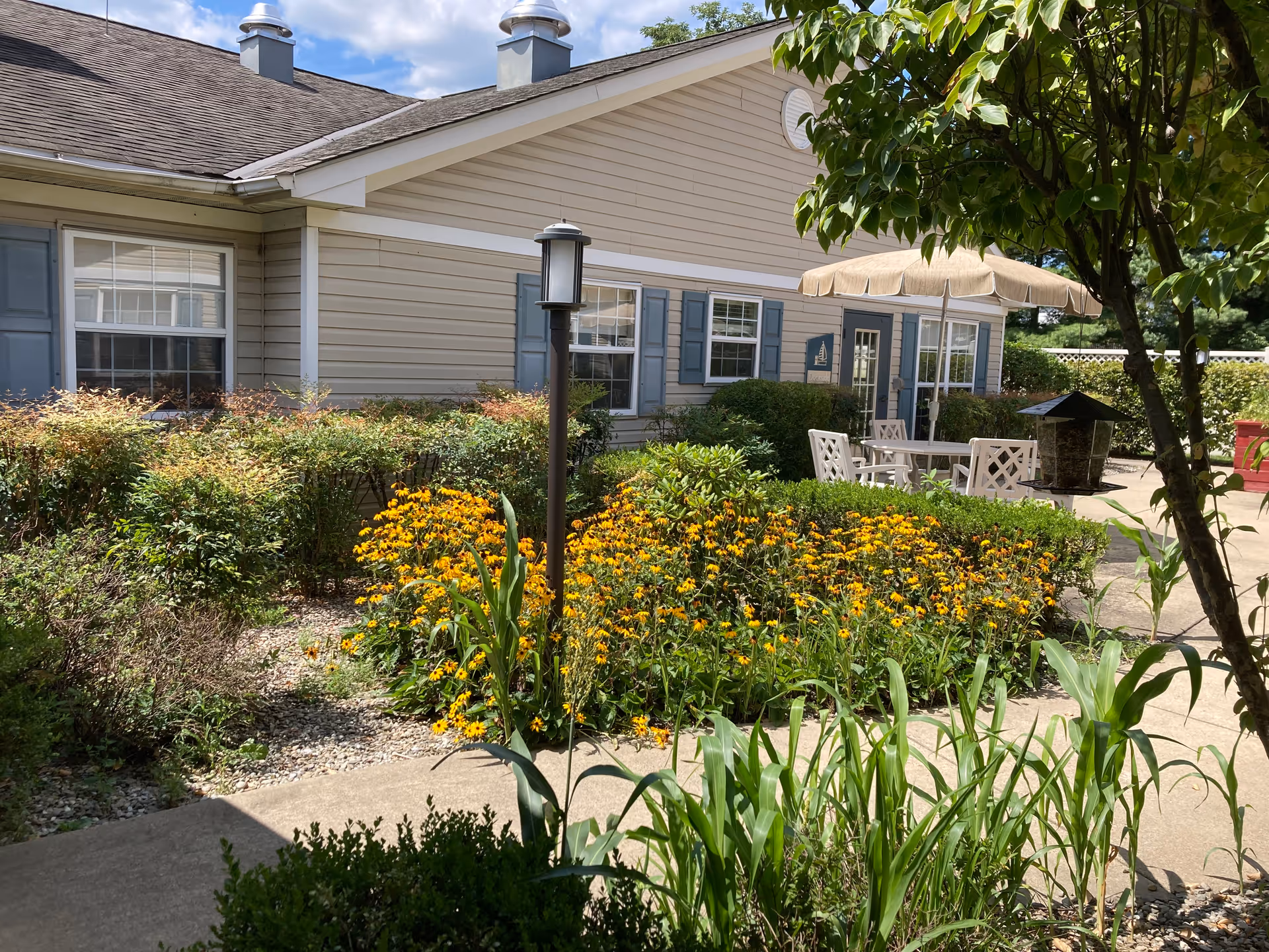 Outdoor garden area at Arden Courts - ProMedica Memory Care Community with yellow flowers, green shrubs, a concrete pathway, a patio table with chairs and an umbrella, and a beige building with blue shutters in the background under a partly cloudy sky.
