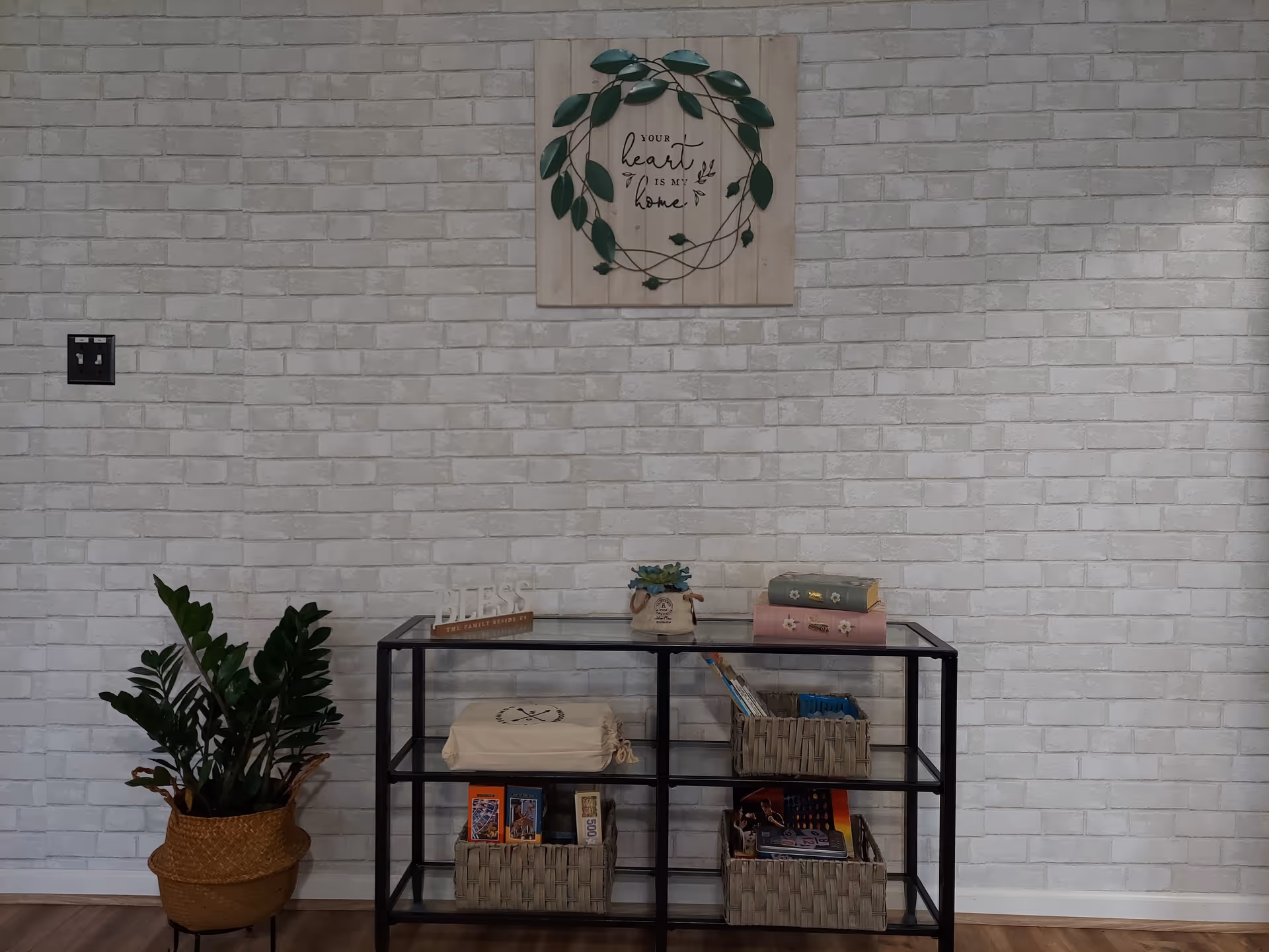 A decorative interior wall with white brick wallpaper, a black metal and glass shelf holding baskets, books, and a small potted plant. A green leafy plant in a woven basket sits on the floor to the left. Above the shelf is a wooden wall art piece with a green leaf wreath and the text 'Your heart is my home.'