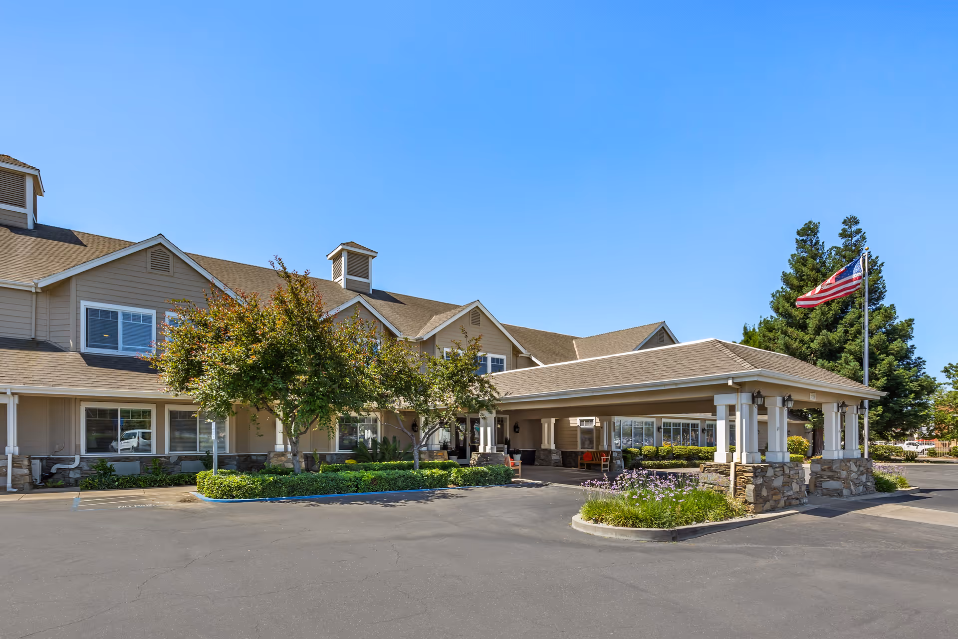 Exterior view of Brookdale Lodi senior living facility showing a beige two-story building with a covered entrance, trees, shrubs, and an American flag on a flagpole against a clear blue sky.