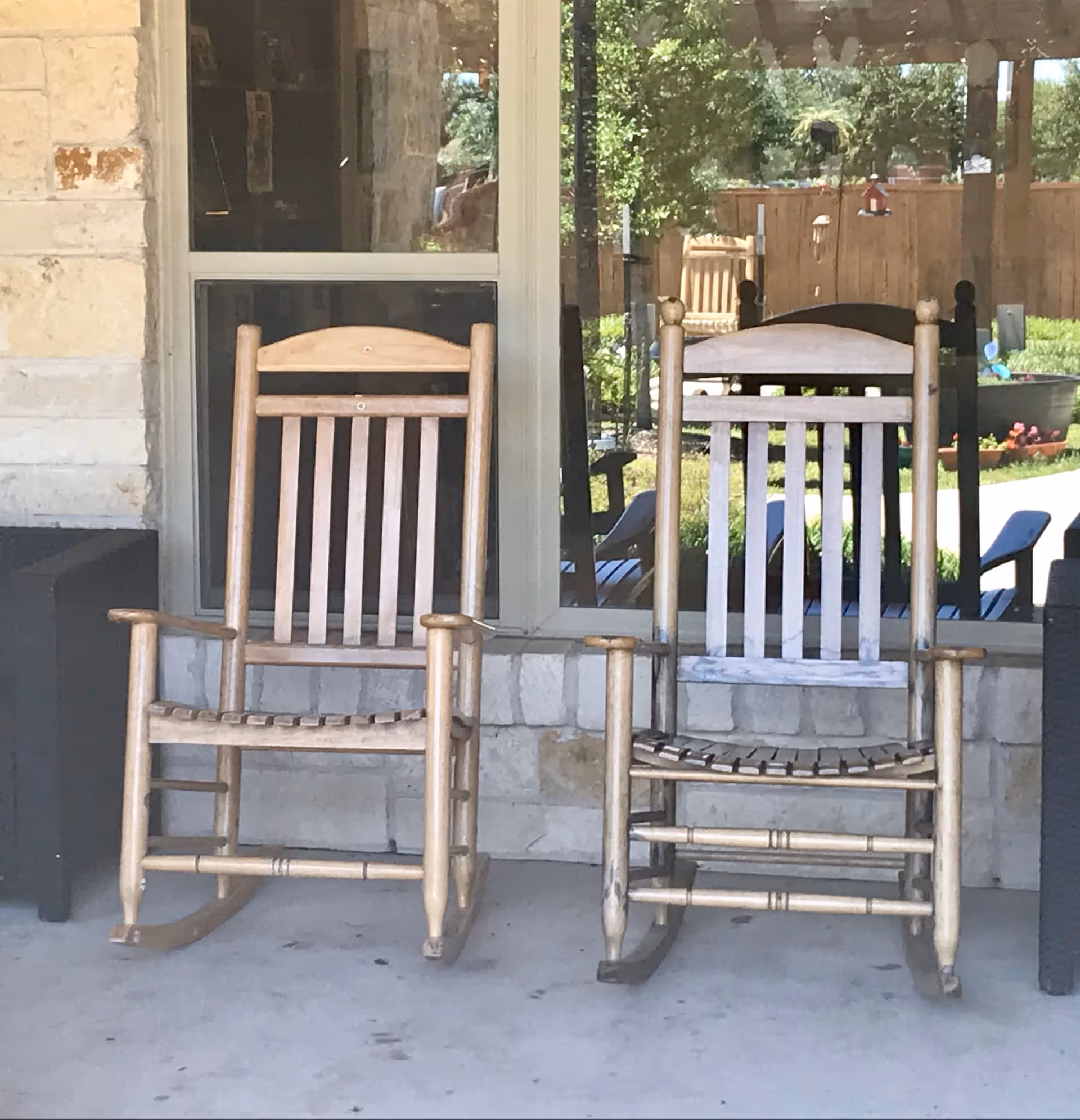 Two wooden rocking chairs placed on a concrete porch in front of a window with a stone wall background. Reflections in the window show greenery and outdoor furniture.