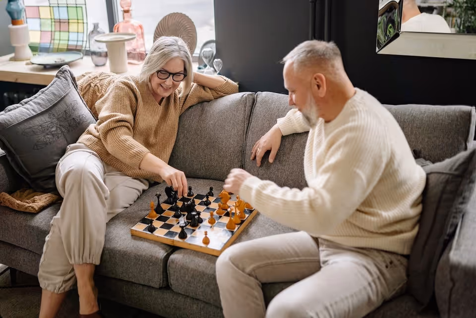An elderly woman and man sitting on a gray couch playing chess together in a cozy living room setting. The woman is smiling and wearing glasses and a brown sweater, while the man is focused on the game wearing a white sweater. Decorative items and a mirror are visible in the background.