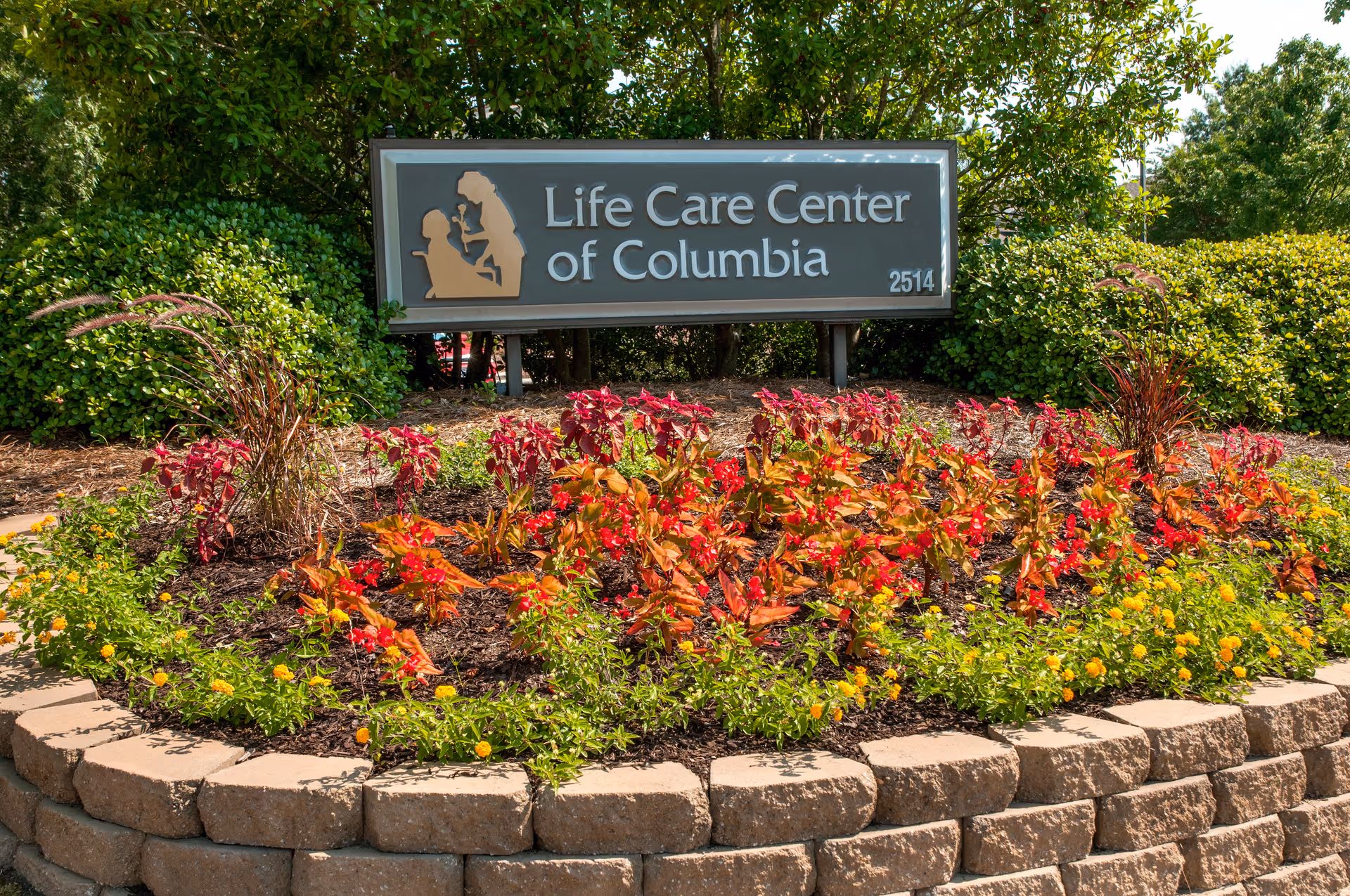 A landscaped flower bed with red, orange, and yellow flowers in front of a sign that reads 'Life Care Center of Columbia 2514', surrounded by green bushes and trees.