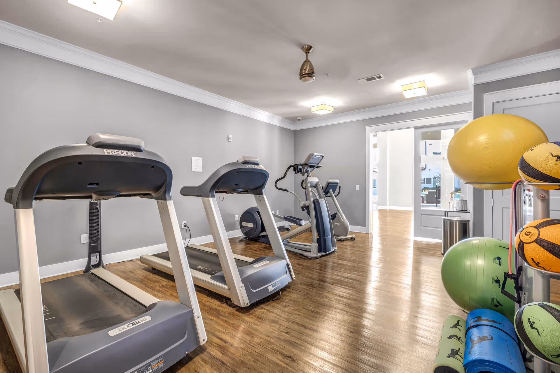 Interior view of a fitness room with two treadmills, an elliptical machine, and various exercise balls and mats neatly arranged on a rack. The room has wooden flooring, gray walls, and ceiling lights with a ceiling fan.