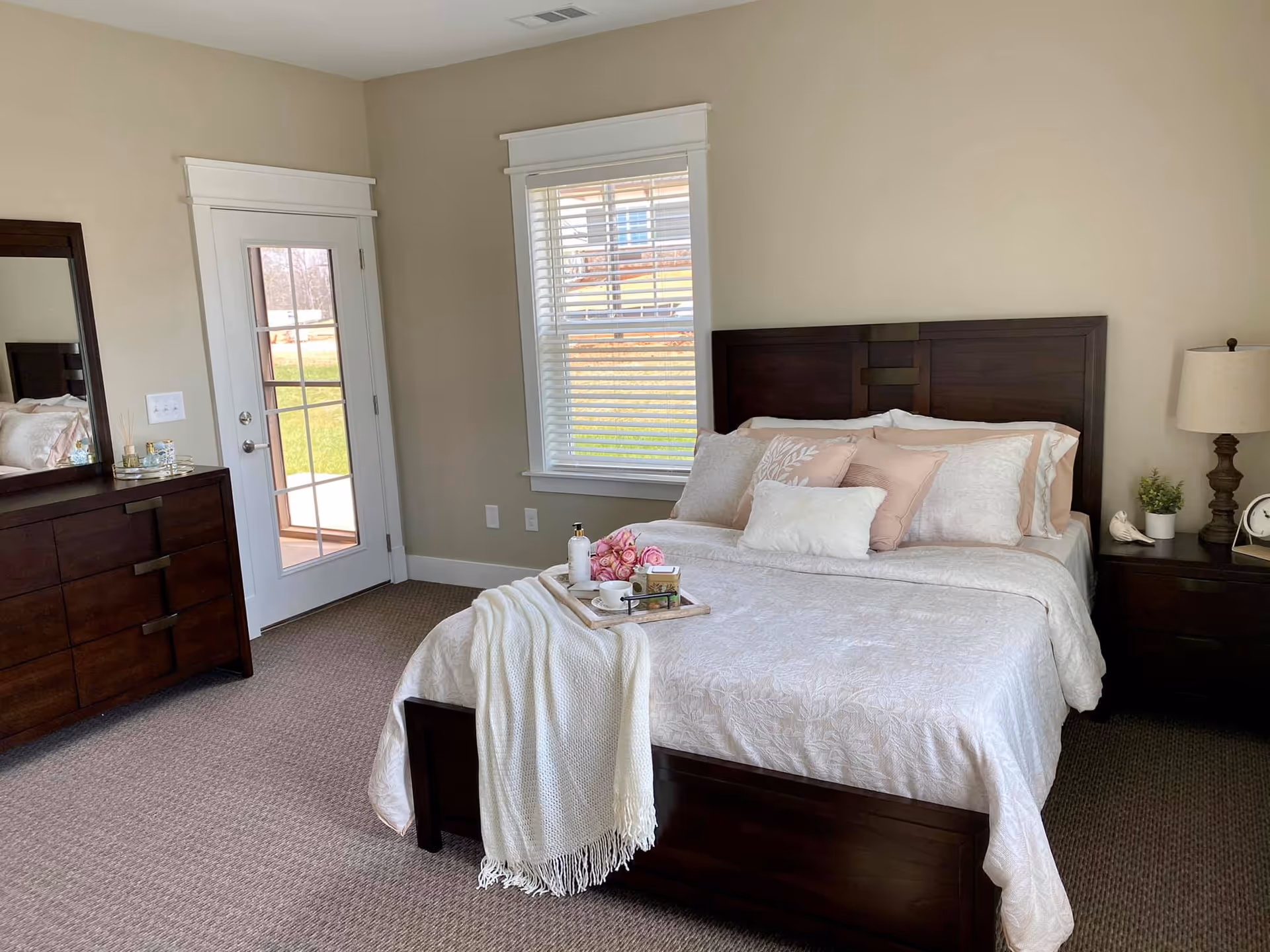A neatly made bed with white and light pink bedding and multiple pillows in a bedroom. A white throw blanket is draped over the foot of the bed with a tray holding a bottle, flowers, and a cup. To the right of the bed is a dark wooden nightstand with a lamp, a small plant, a decorative bird, and a clock. To the left is a dark wooden dresser with a mirror above it. There is a window with white blinds and a glass door leading outside, with beige walls and carpeted floor.