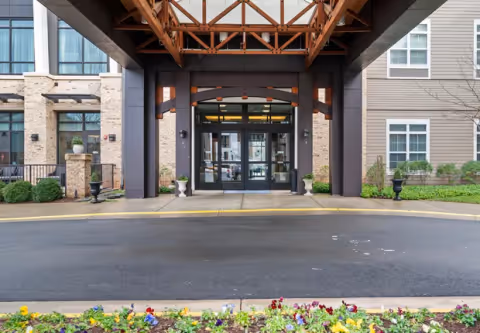 Covered main entrance of a residential building with a timber canopy, glass double doors, and a flowerbed in the foreground.