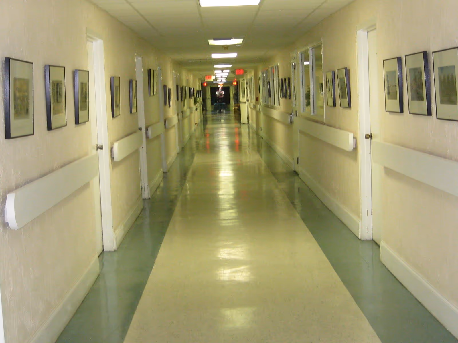 A long, empty hallway in a facility with light-colored walls and a shiny floor. The hallway is lined with closed doors on both sides and handrails along the walls. Framed pictures are hung evenly along both walls. The ceiling has fluorescent lights, and exit signs are visible at the far end.