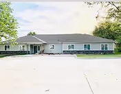 Single-story building with a gray exterior and stone accents along the bottom, surrounded by trees and a large concrete driveway in front under a partly cloudy sky.