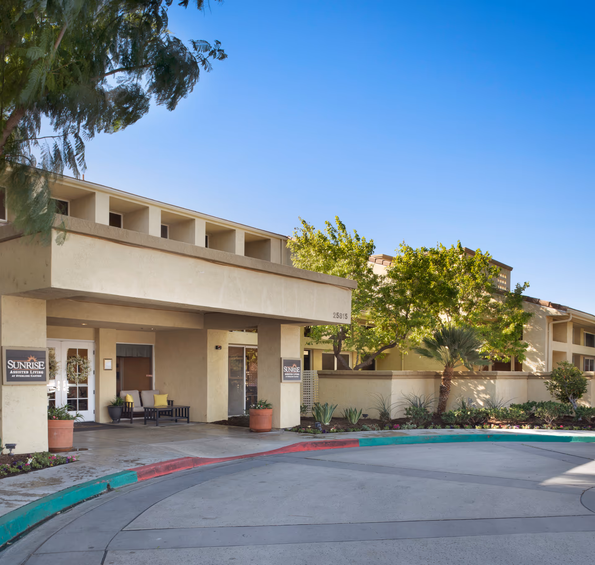 Exterior view of Sunrise At Sterling Canyon assisted living facility entrance with a covered drop-off area, potted plants, seating, and trees under a clear blue sky.