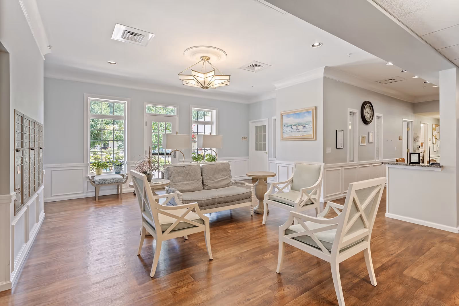 A bright and spacious common area with wooden flooring, white walls, and large windows letting in natural light. The room is furnished with a beige cushioned sofa, four white wooden chairs with light cushions, two round wooden side tables, and two table lamps. There is a painting on the wall and a clock further down the hallway. The area appears clean and inviting, suitable for socializing or relaxing.