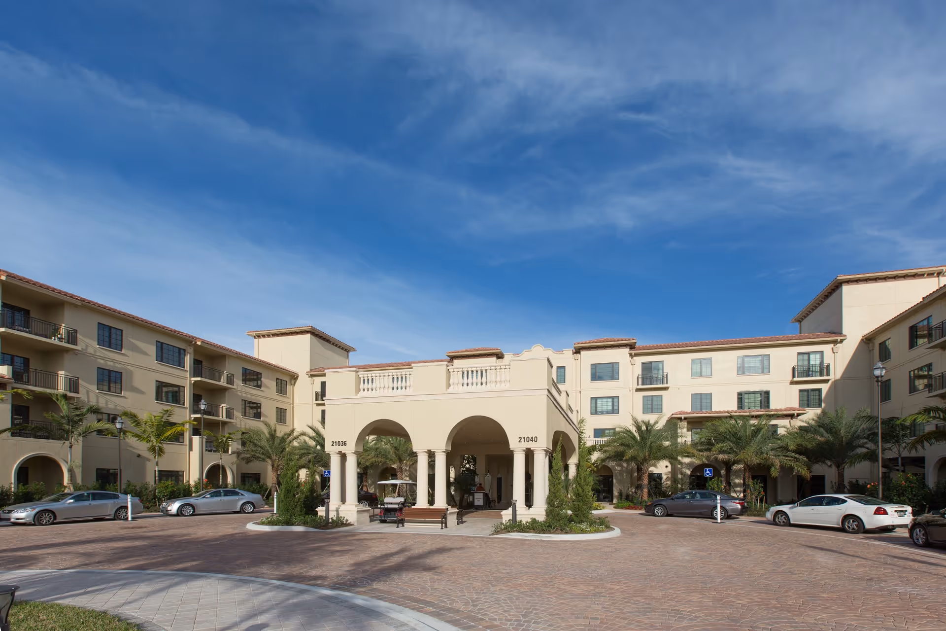 Front entrance and circular driveway of a Mediterranean-style senior living facility with palm trees and parked cars under a blue sky.
