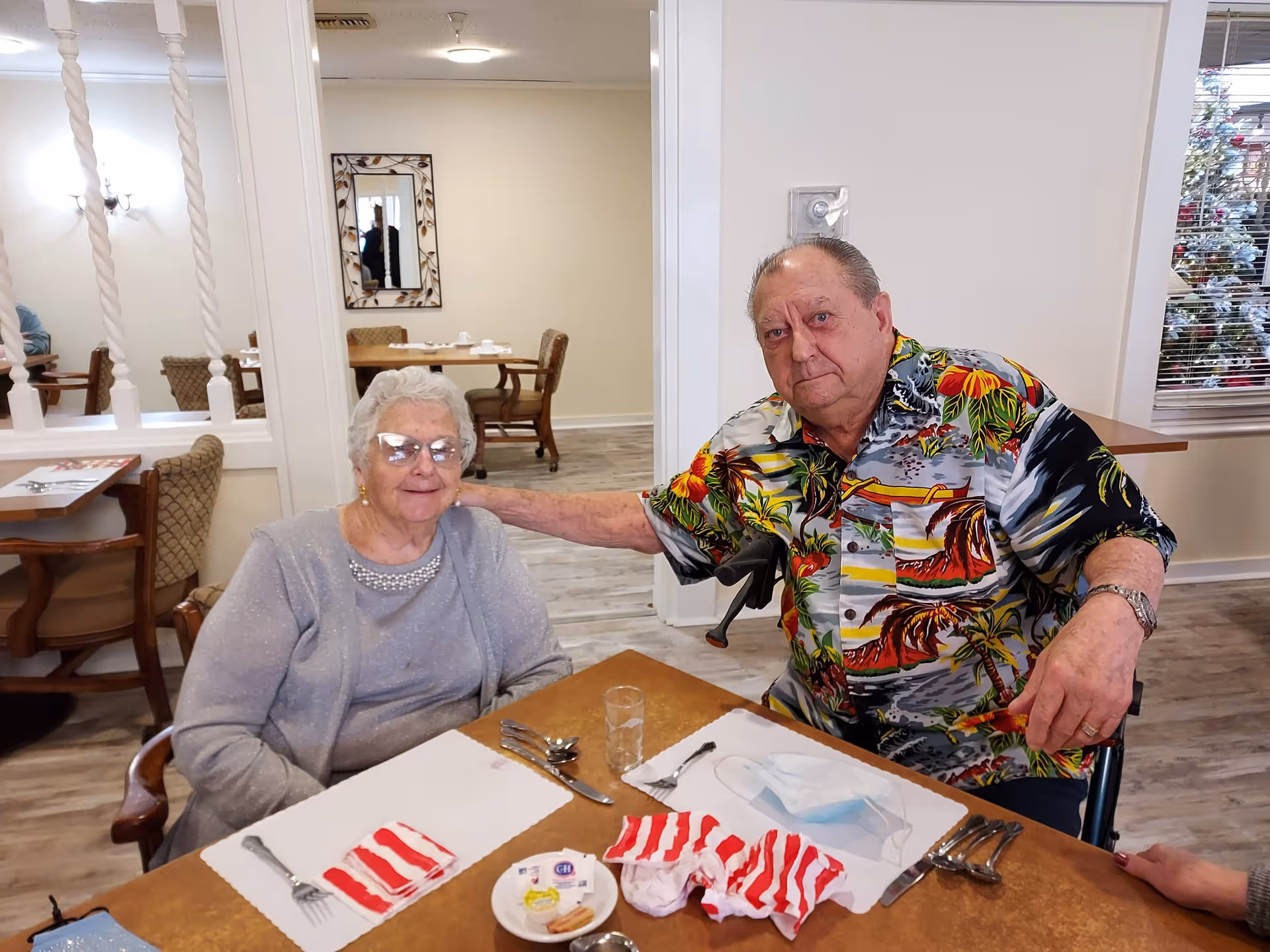 An elderly woman and an elderly man sitting together at a dining table in a retirement community dining room. The woman is wearing glasses and a light gray sweater, while the man is wearing a colorful tropical shirt and has his arm around the woman. The table is set with placemats, utensils, a glass, and some small plates with food items. The background shows other dining tables and chairs, a mirror on the wall, and a window with blinds.