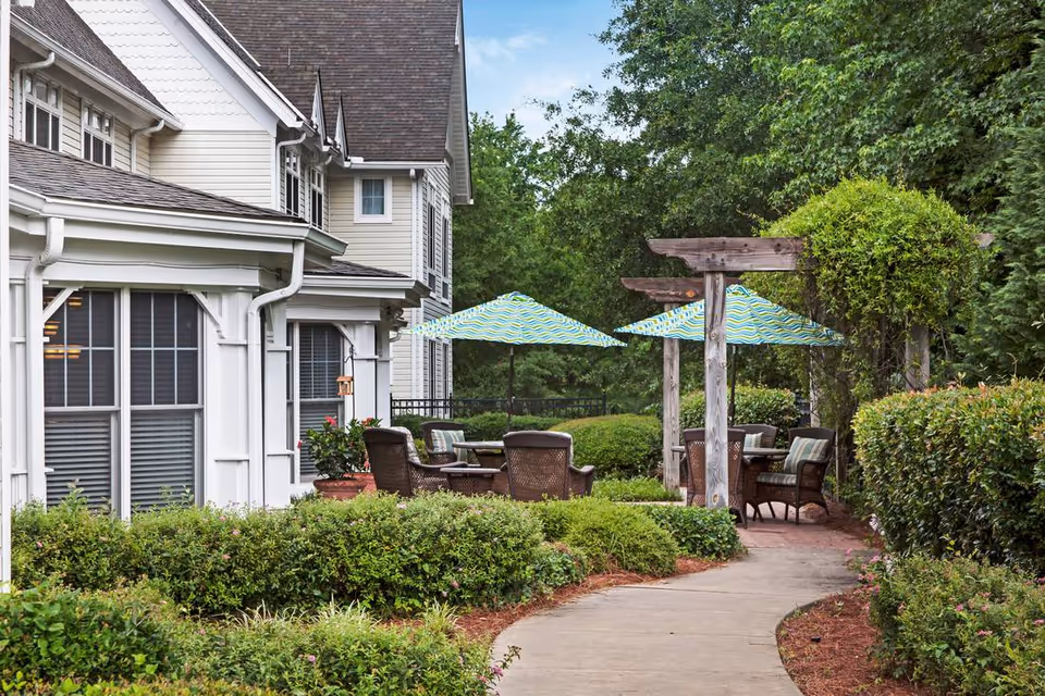 Outdoor seating area at Assisted Living-Five Forks featuring wicker chairs and tables under blue and green striped umbrellas, surrounded by lush green bushes and trees along a curved concrete pathway next to a light-colored building.