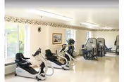 A bright exercise room with large windows letting in natural light. The room contains several pieces of fitness equipment including recumbent exercise bikes and weight machines. The walls are light-colored with a decorative border near the ceiling, and framed artwork is visible on the wall.