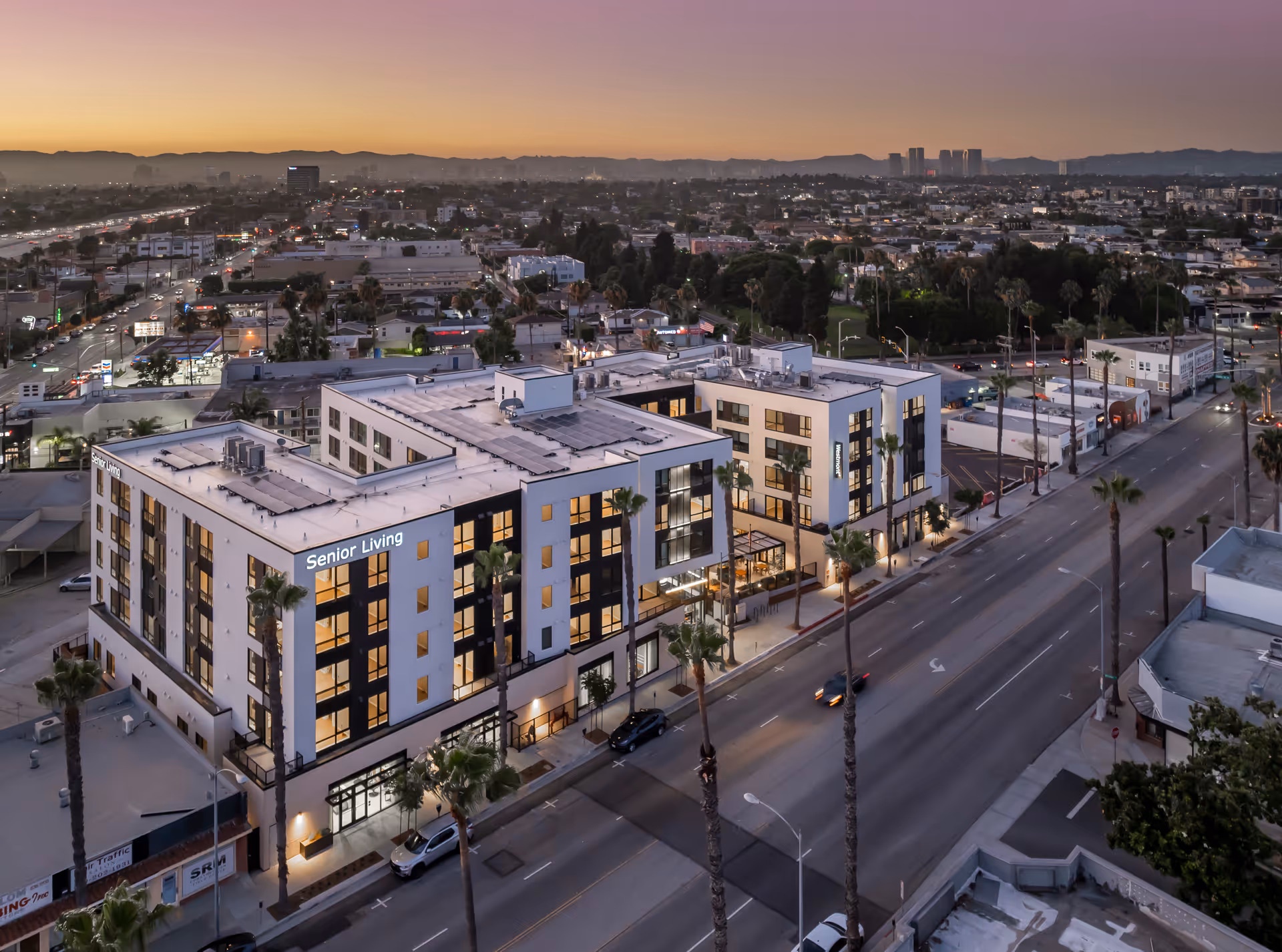 Aerial view of a modern senior living facility named Westmont of Culver City at dusk, showing a multi-story building with illuminated windows, palm trees lining the street, and a cityscape with mountains in the background.