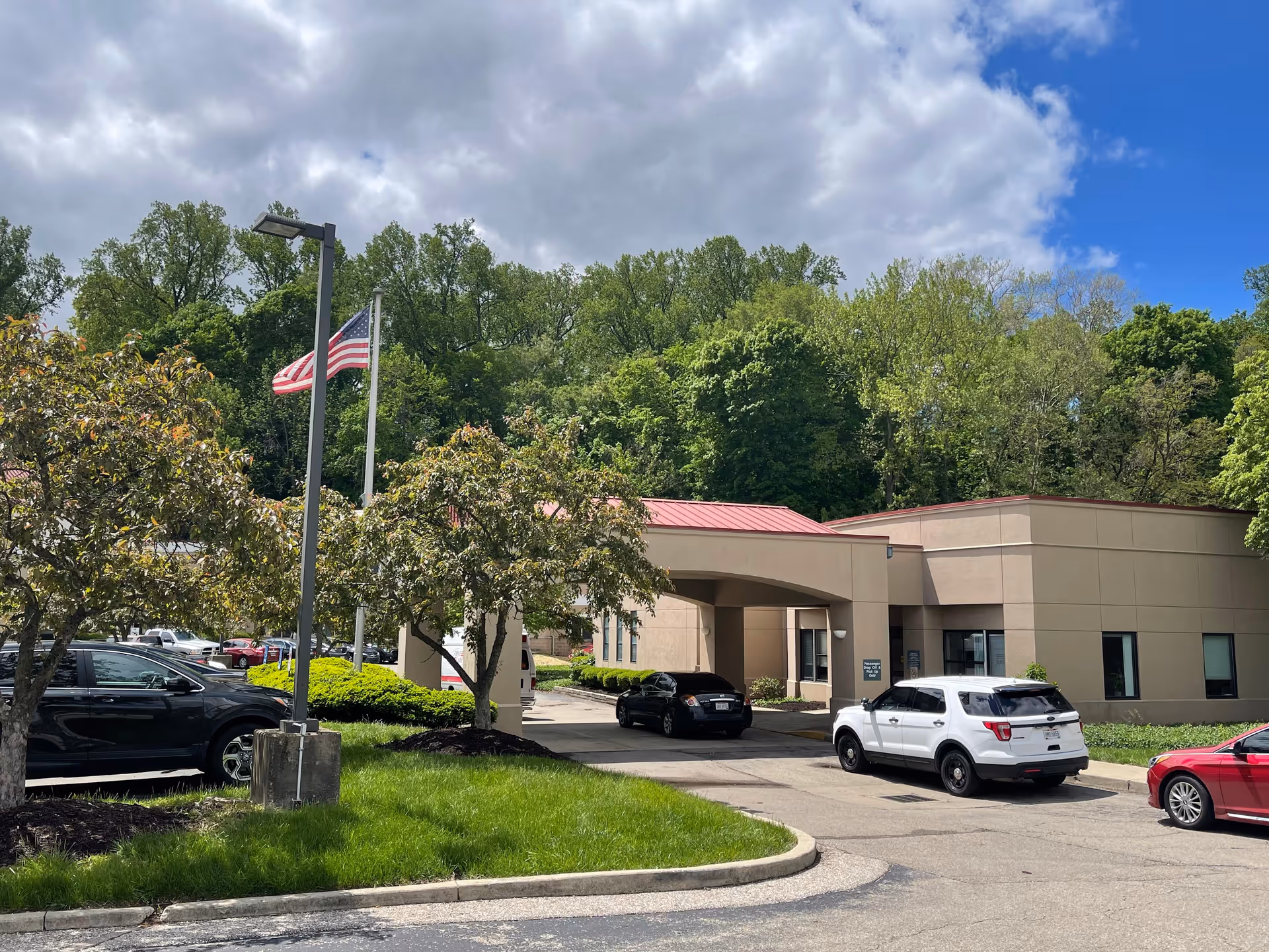 Exterior view of a single-story building with a covered entrance and several parked cars. There are trees and greenery surrounding the building, an American flag on a pole, and a partly cloudy sky overhead.