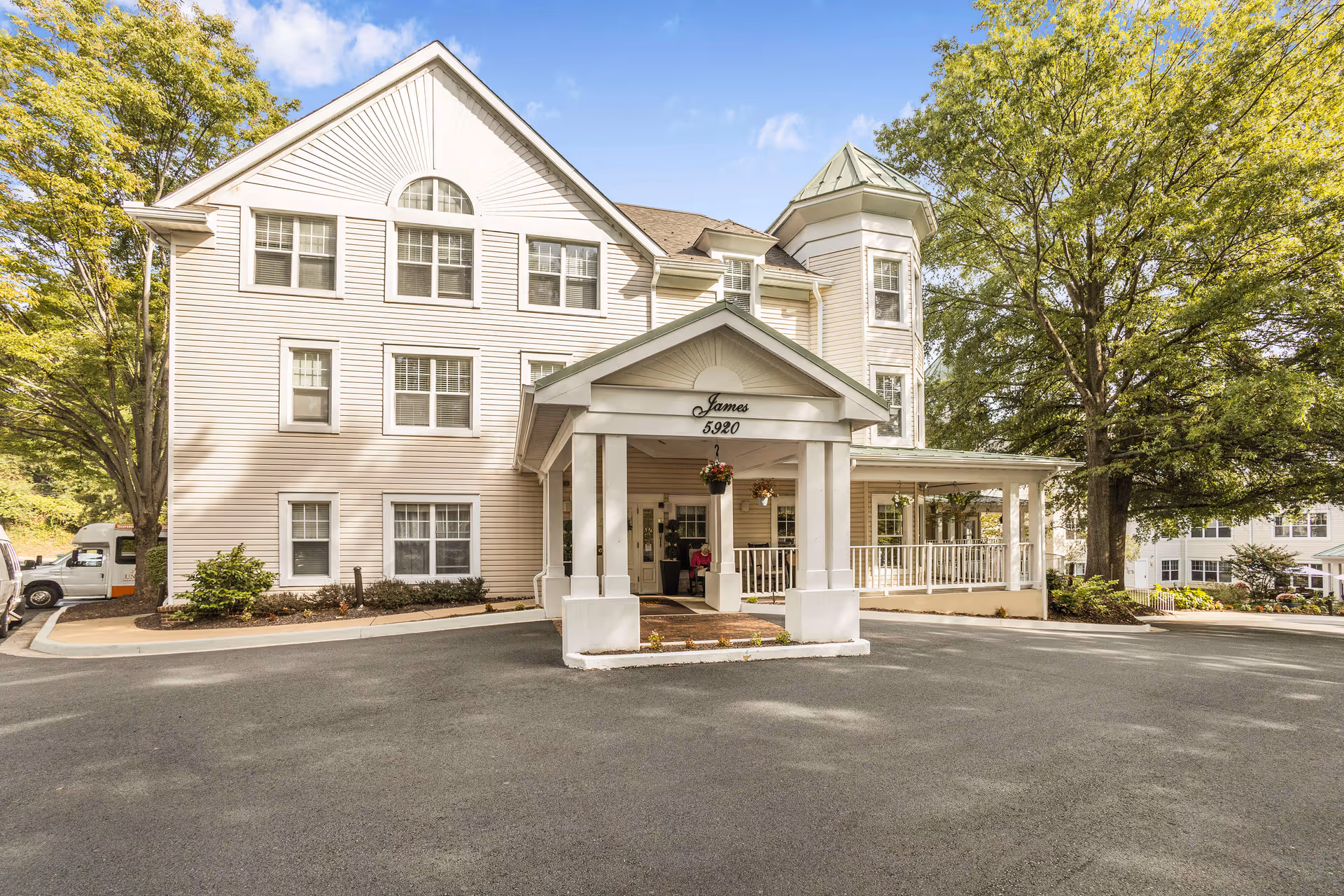 Exterior view of a three-story senior living facility building with white siding, multiple windows, and a covered entrance with the name 'James' and the number '5920' above it. There are trees and landscaping around the building and a paved driveway in front.