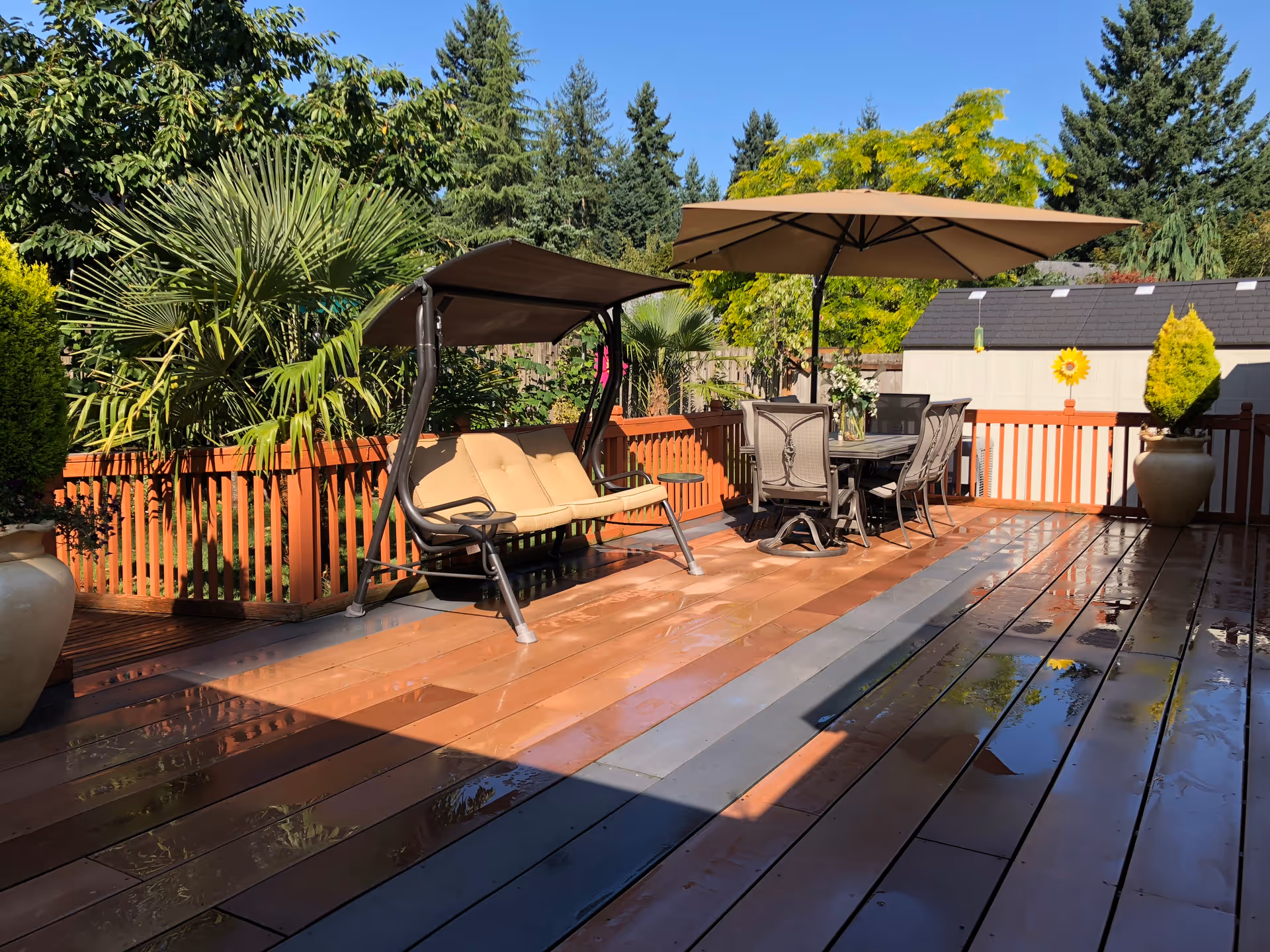 Outdoor wooden deck area with a cushioned swing bench on the left and a dining table with six chairs under a large umbrella on the right. The deck is surrounded by a wooden fence and lush green trees and plants. There are large potted plants on either side of the deck, and the deck surface is wet with some reflections visible.