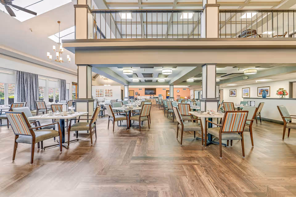 Spacious two-story dining room with multiple set tables and chairs, large windows and overhead lighting in a senior living facility.