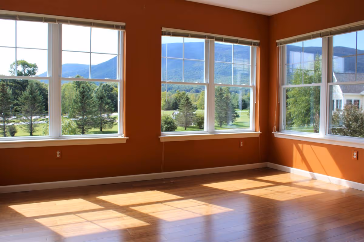 Empty room with large windows showing a scenic view of trees and mountains outside. The walls are painted orange and the floor is wooden with sunlight streaming in through the windows.