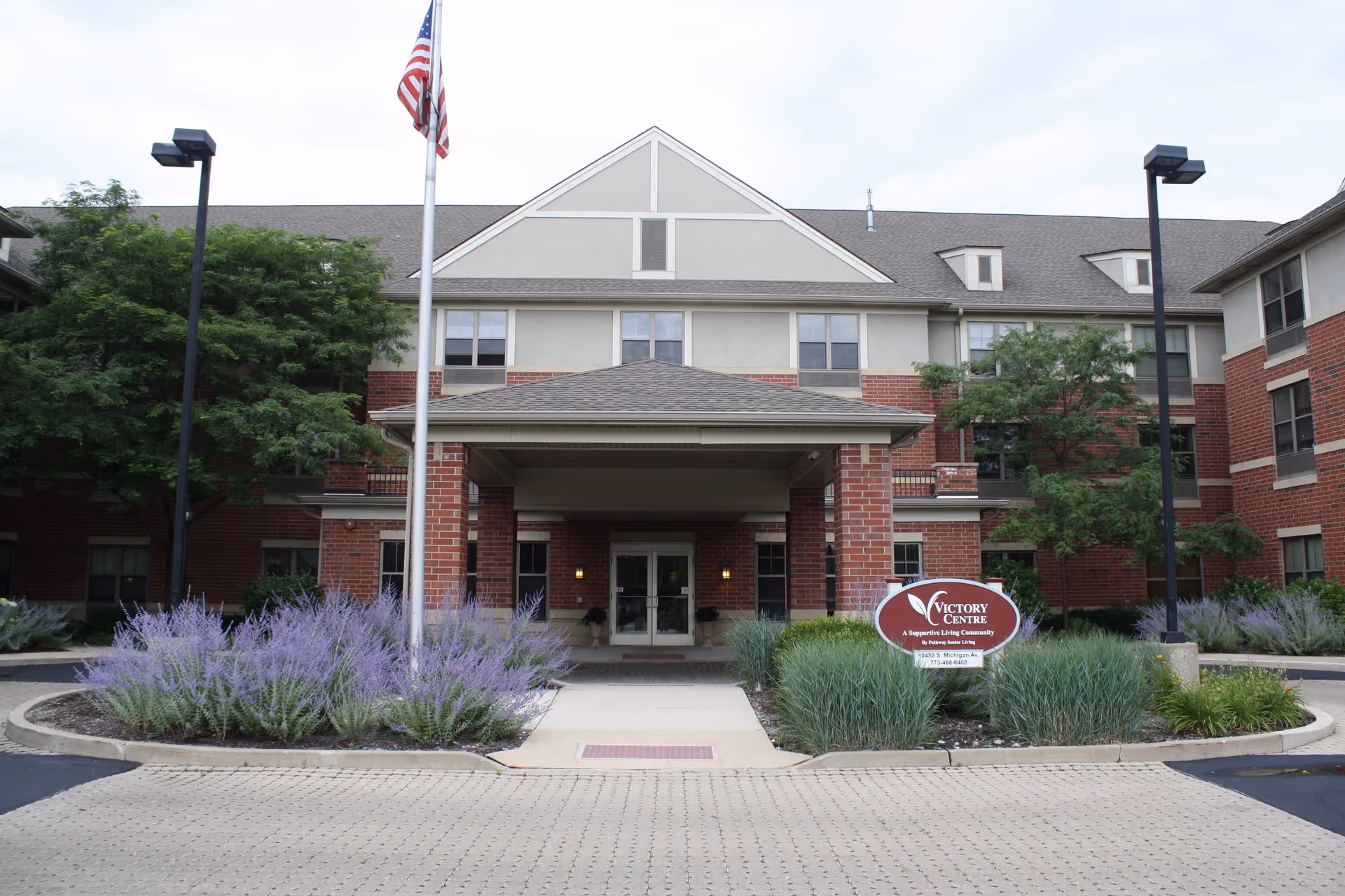 Front exterior view of Victory Centre of Michigan Ave - Roseland Supportive Living & Memory Care building with a covered entrance, American flag on a flagpole, landscaped bushes with purple flowers, and a sign displaying the facility's name and contact information.