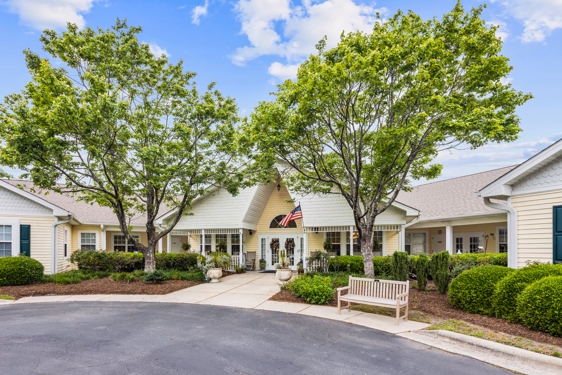 Exterior view of TerraBella Southport facility showing a single-story building with light yellow siding, white trim, and a gray roof. There are two large green trees and well-maintained bushes in front of the entrance. An American flag is displayed above the double glass doors. A wooden bench is placed near the sidewalk leading to the entrance under a partly cloudy blue sky.