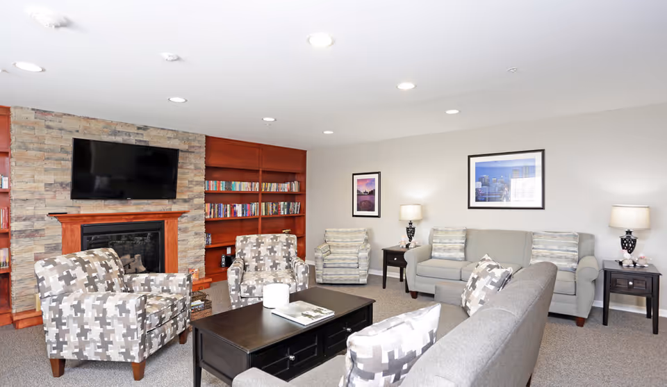 Bright communal living room with sofas, patterned armchairs, a stone fireplace, wall-mounted TV and built-in bookshelves.
