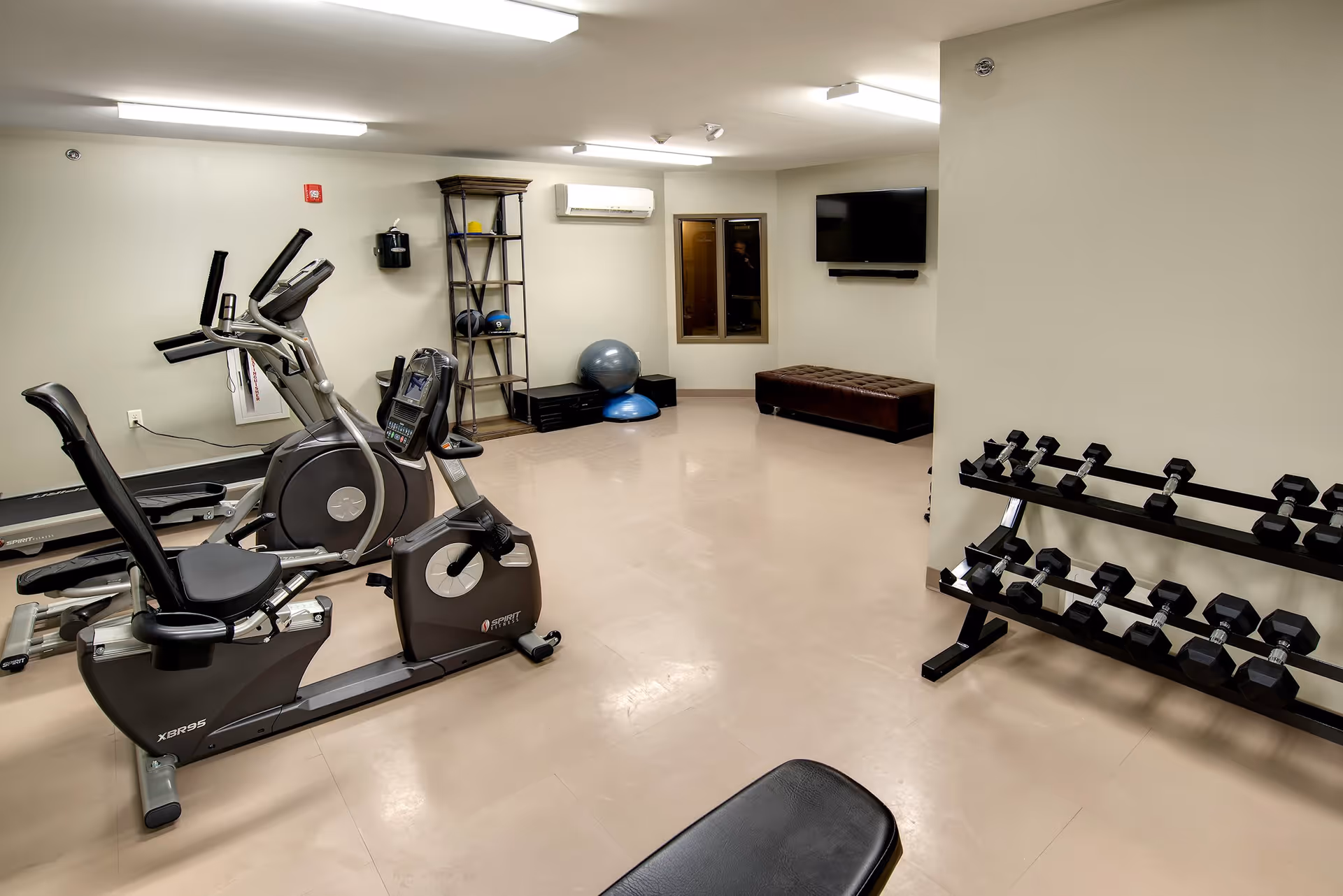 A fitness room with exercise equipment including a recumbent bike, elliptical machine, dumbbell rack, exercise balls, and a wall-mounted TV. The room has beige walls, a tiled floor, and fluorescent ceiling lights.