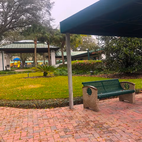 Outdoor area at Weinberg Village Assisted Living featuring a green metal bench on a brick-paved path, a covered structure with a green roof, palm trees, manicured grass, and various shrubs and plants.