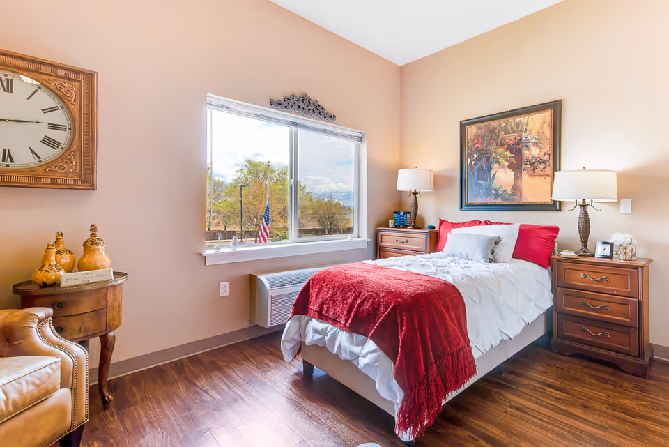 A bright furnished bedroom with a single bed dressed in white and red linens, two wooden nightstands with lamps, a large window, and decorative wall art.