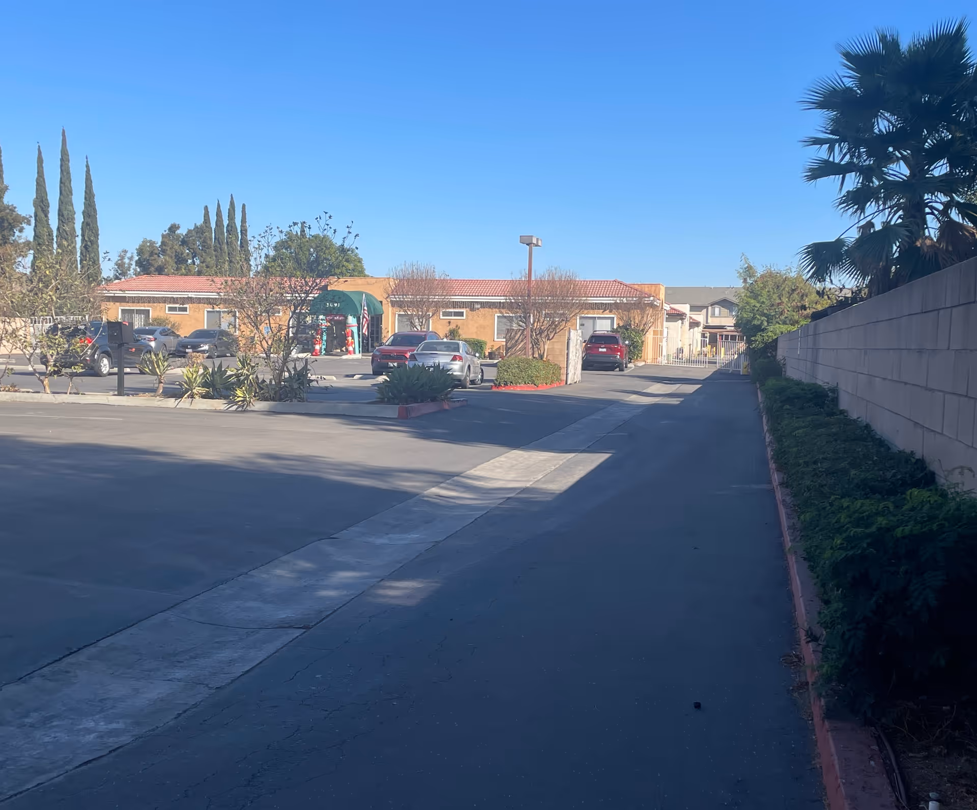 View of the parking lot and entrance of CalOaks Senior Living facility on a clear sunny day, with several parked cars, a building with a red tile roof, green awning, and some trees and shrubs around the area.