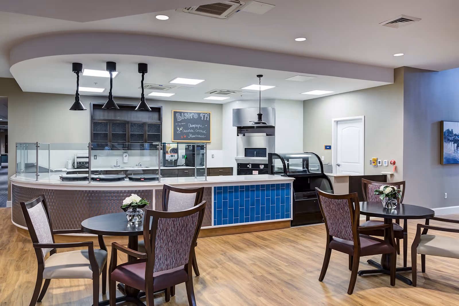 A modern dining area with round tables and chairs, featuring a serving counter with glass partitions and kitchen appliances in the background. The floor is wooden, and the ceiling has recessed lighting and hanging pendant lights. There is a chalkboard menu on the wall behind the counter.