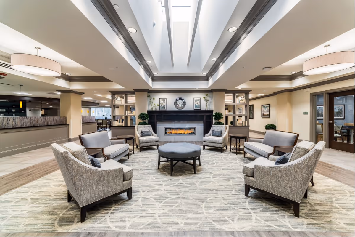 A spacious and well-lit senior living facility common area with a modern fireplace centered on the far wall. The room features a large skylight, recessed lighting, and neutral-toned walls. Comfortable seating includes two armchairs and two loveseats arranged around a round ottoman on a patterned area rug. Decorative plants and framed artwork flank the fireplace, and there are open shelving units on either side. The area extends into other parts of the facility with visible dining and seating spaces.